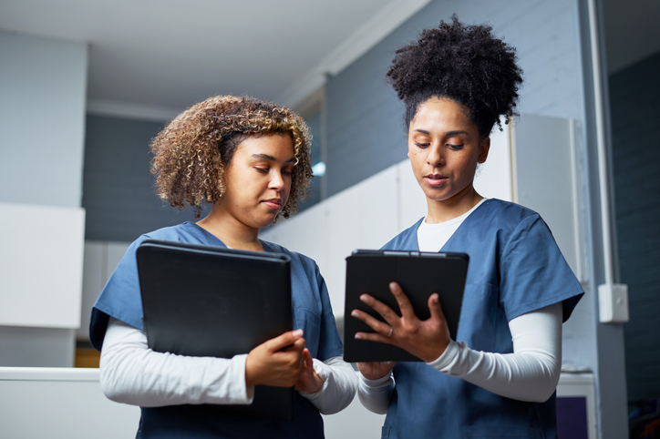 Two nurses wearing professional uniforms review and exchange medical data on a digital tablet. 
