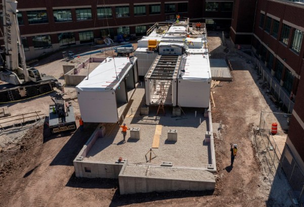 Multiple pre-fabricated buildings placed together on concrete foundation. One building is being lowered by crane. All buildings and foundation are at a construction site near an existing commercial building.