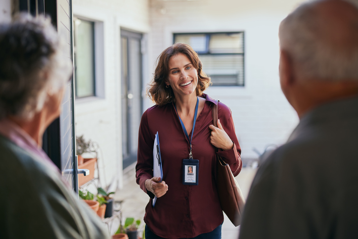 Elder couple open the door and meeting friendly smiling social worker visiting.