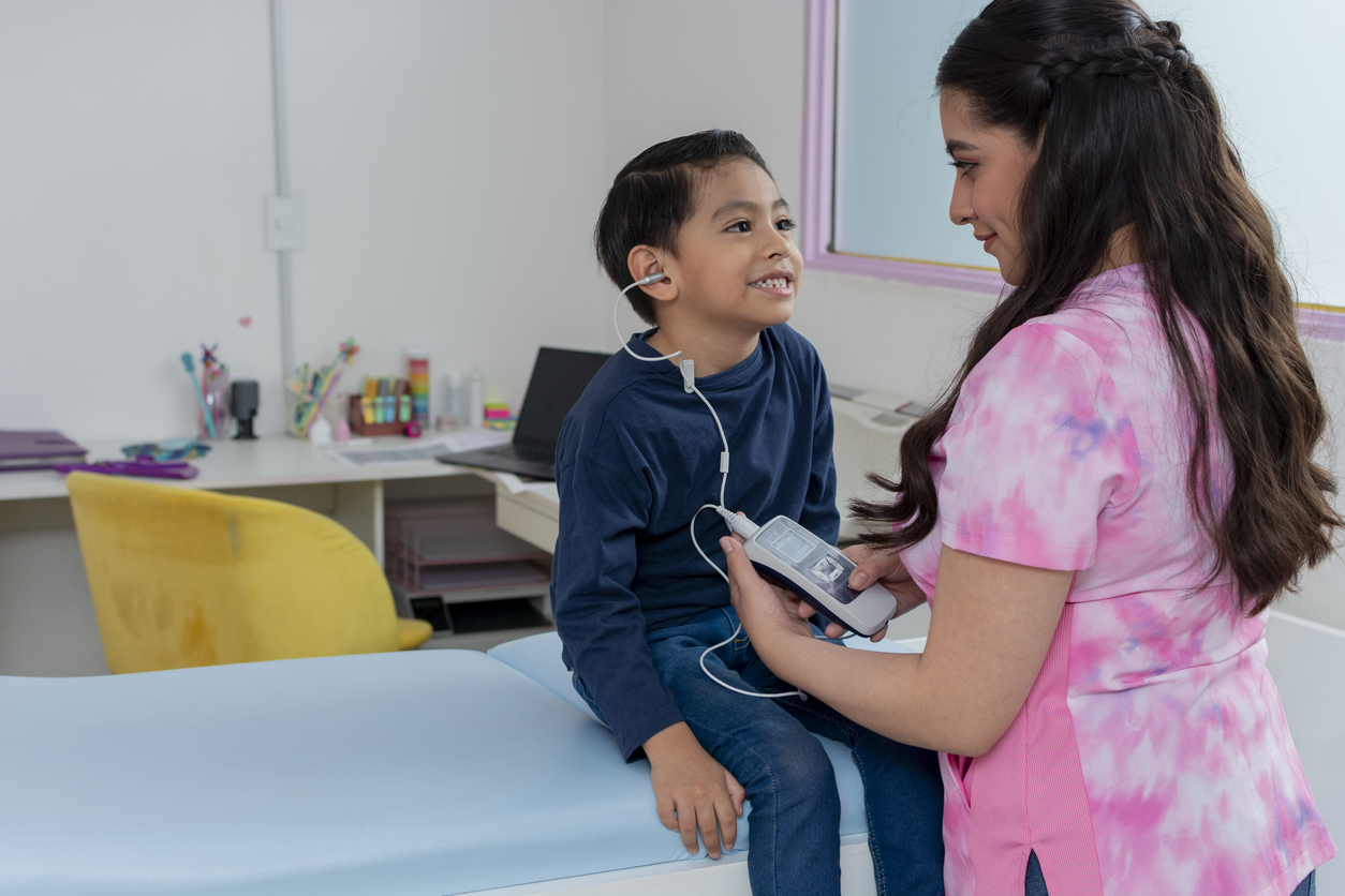 Young boy sits on examination table while a female audiologist performs a hearing test in a doctor's office. 