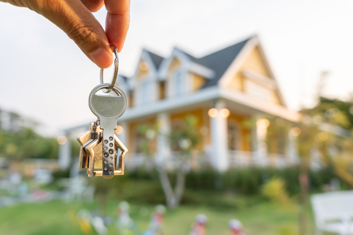 Real estate agent holding key for new owner. Keys are in focus, with yellow house with wide wraparound porch supported by white pillars in the background.

