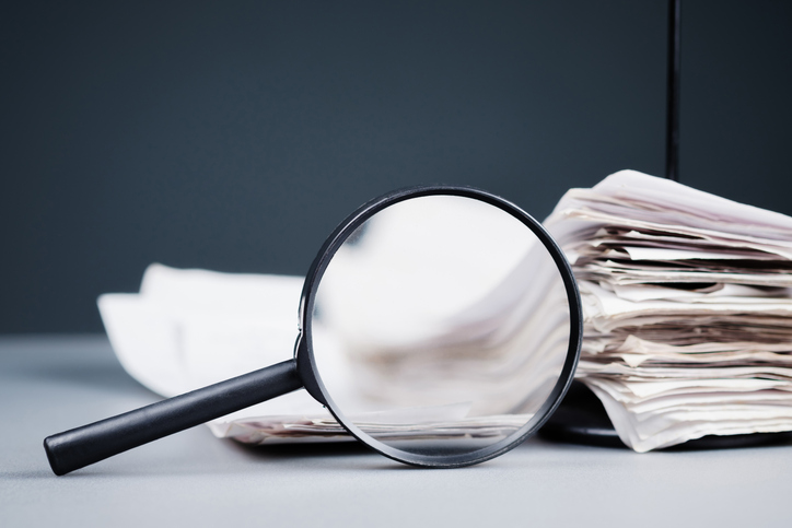 A magnifying glass with a stack of papers on the desk needing to be inspected. 