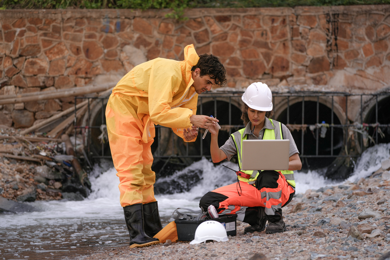 Two environmental field workers in protective gear conduct a water pollution inspection at a drainage outlet, collecting samples and analyzing data for contamination levels.
