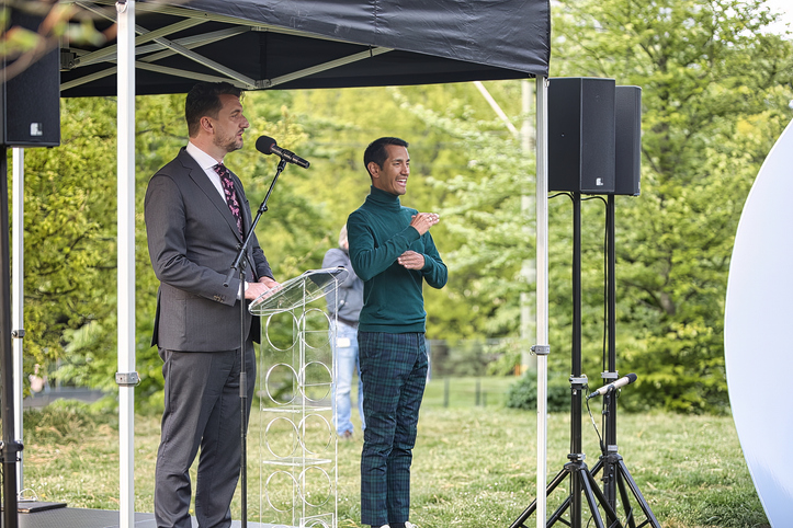 A man in suit is giving an outdoor speech with live sign language interpreter standing to the right.