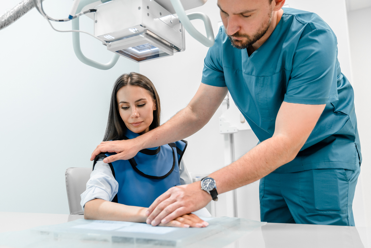 Male healthcare worker works with an x-ray machine in a modern hospital while patient sitting as he adjusts her arm for x-ray photo.