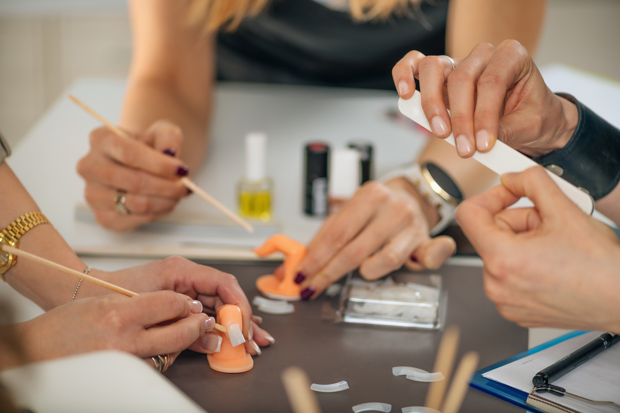 Training for Nail Technician – Manicurist. One person filing their nails with an emery board and two people working on applying nails to orange freestanding practice fingers.