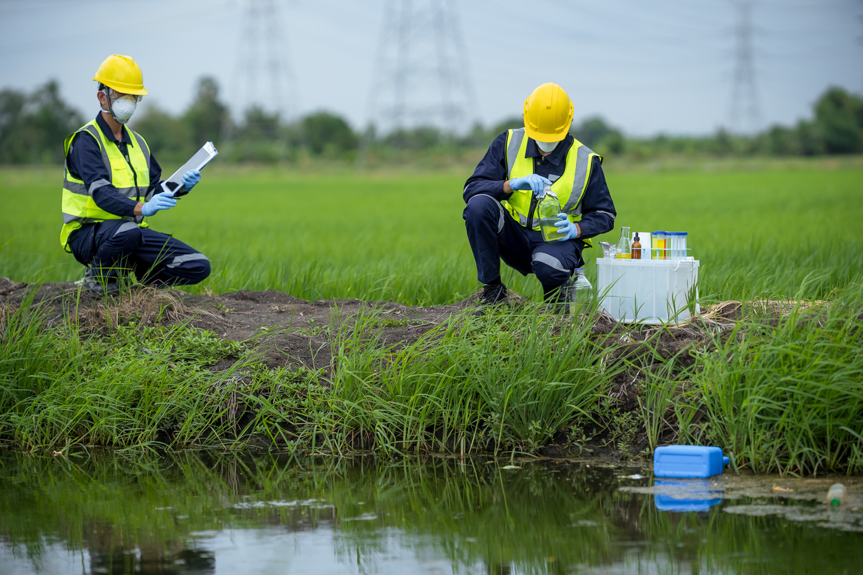 Two scientists with tablet and soil sample collection kit collaborate on research. Both are crouching on dirt next to grass and a small body of water.