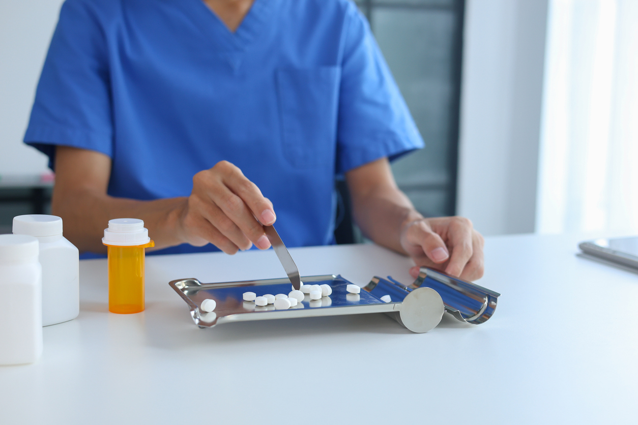 Person wearing scrubs Counting White Medicine Pills on Stainless Steel Counting Tray in Pharmacy.

