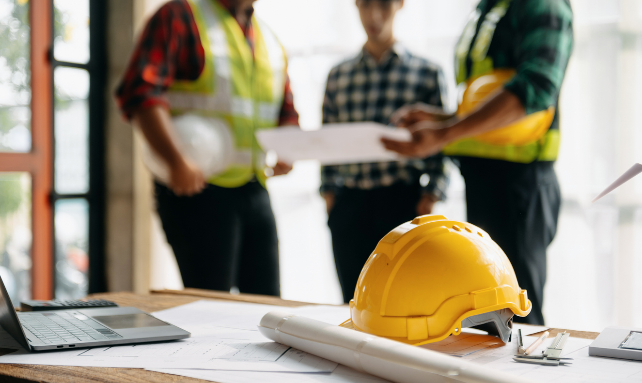 Construction team in background wearing hardhats on construction site. Table with blueprints and laptop. 