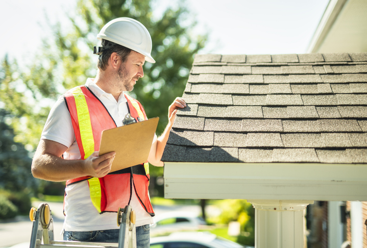 A man with hard hat standing on steps inspecting house roof.