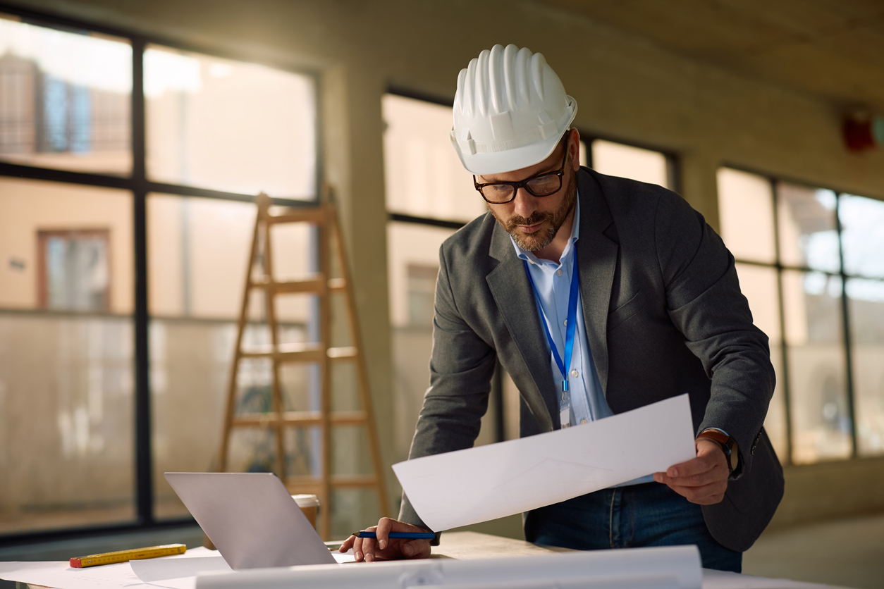 Male architect examining blueprints while working on laptop at construction site.

