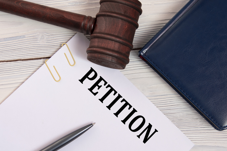 Petition image featuring a gavel, paper clips and the word petition on a piece of pager on a desk next to a blue notebook. 