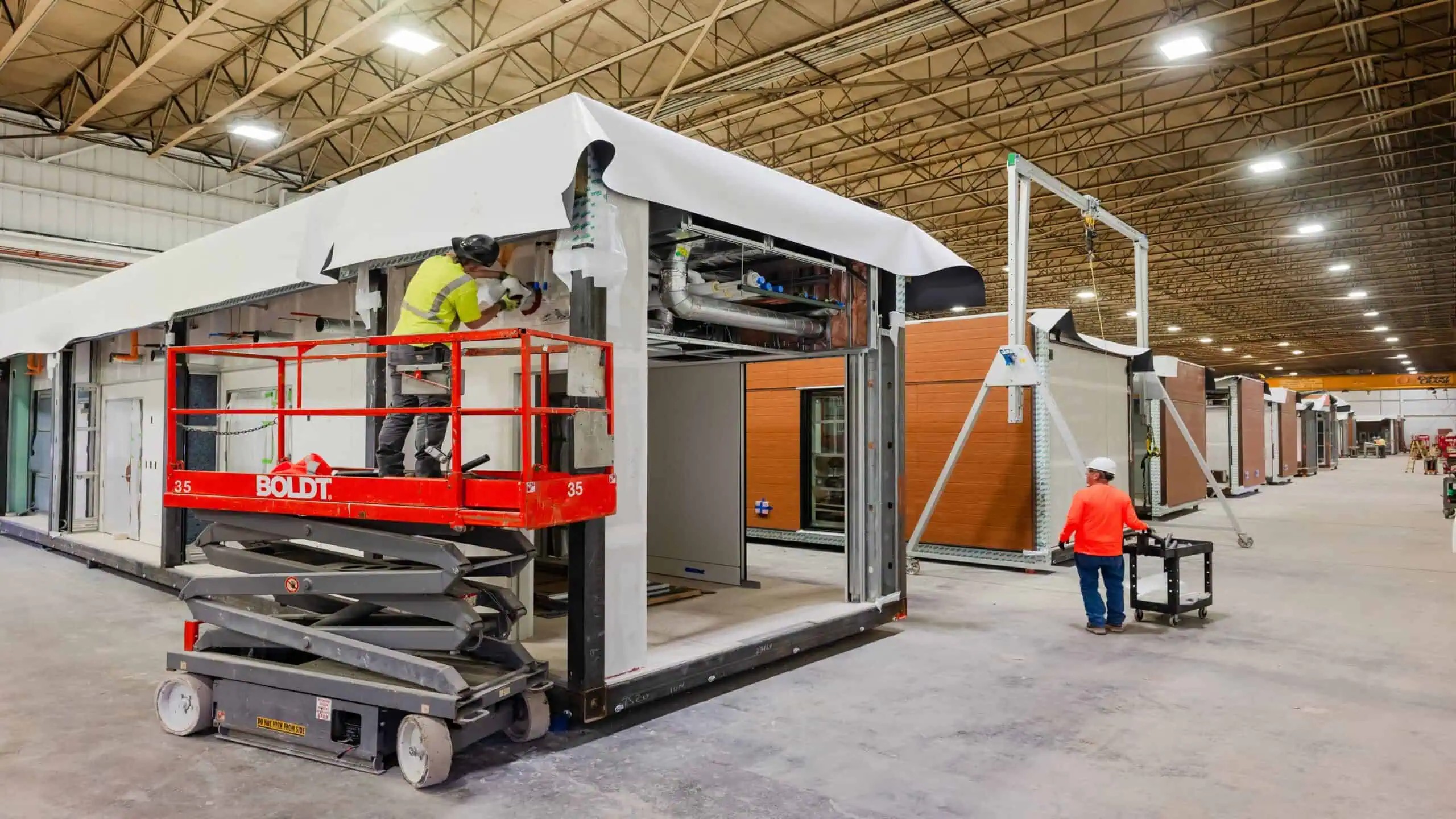 Individual in safety shirt on a scissor lift working on a pre-fabricated building in a factory dedicated to constructing pre-fabricated buildings. Another individual is standing on the concrete floor of the factory with a wheeled cart.