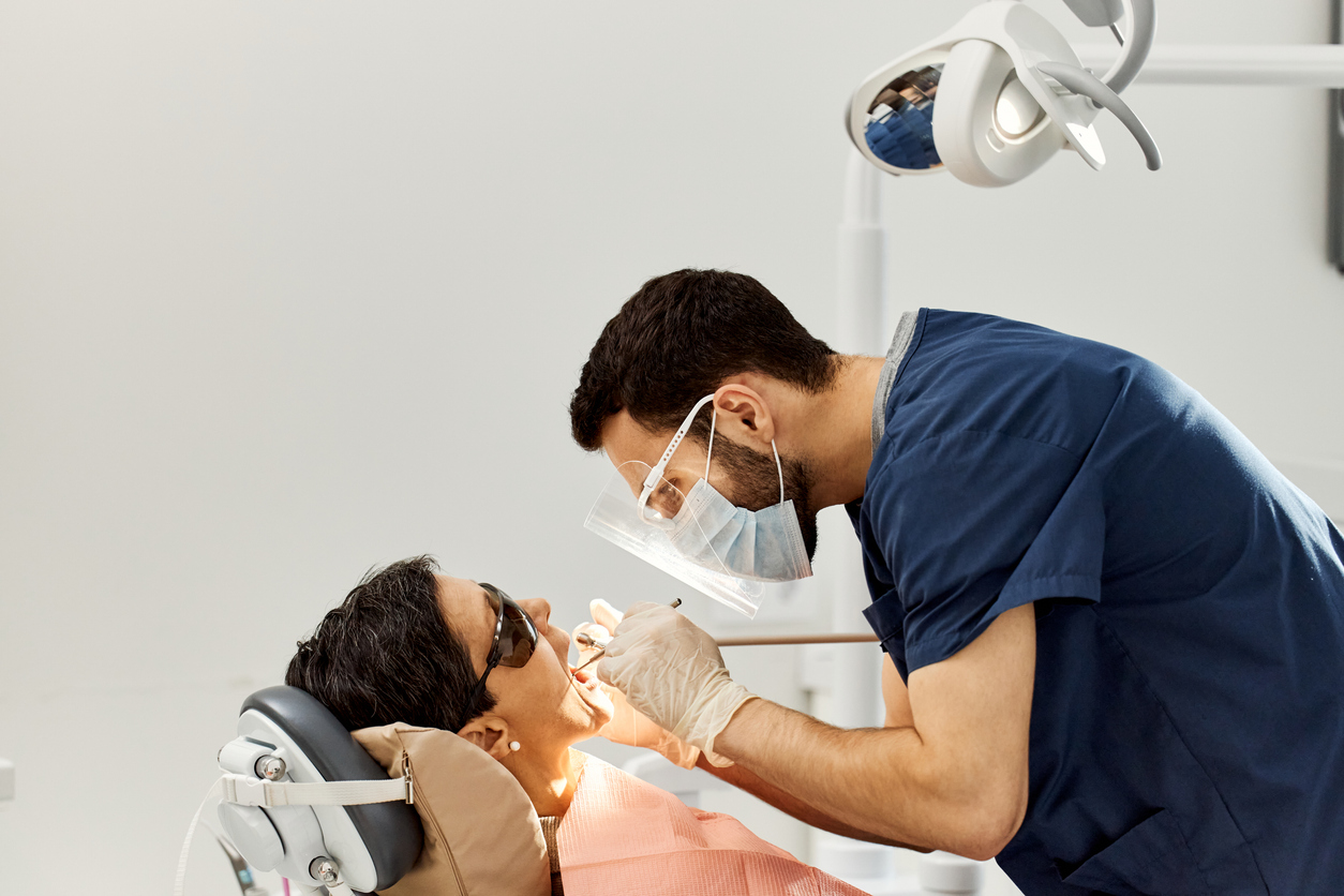 Middle aged Caucasian woman sitting in dental chair, receiving oral examination from young adult Middle Eastern dentist wearing mask and gloves in modern dental office
