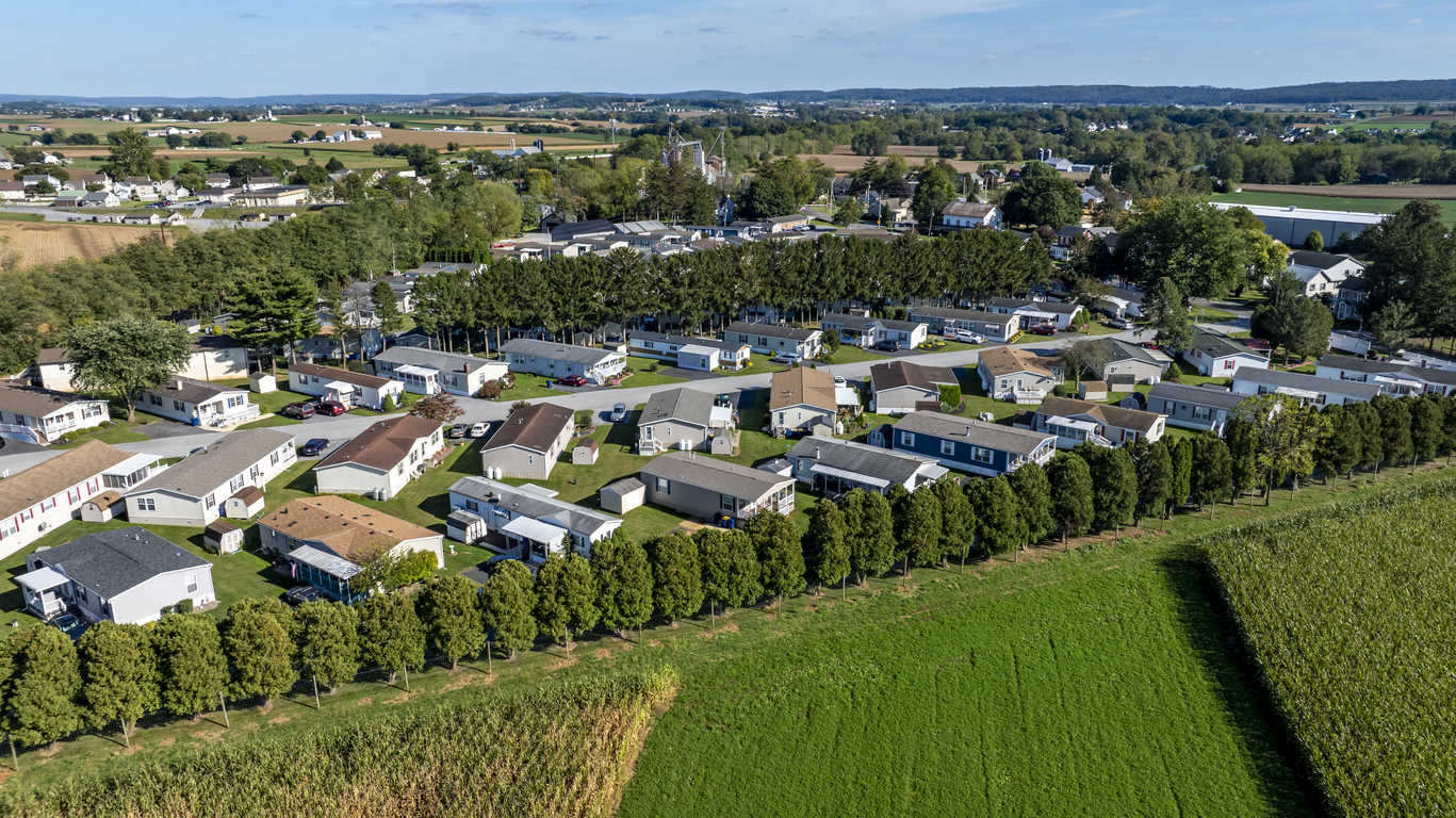 A collection of mobile, manufactured homes aligned in rows in a rural community. The houses are arranged in a neat pattern with trees and open fields nearby. Farms are visible in the distance.

