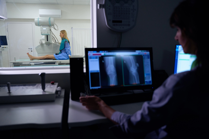 Woman sitting on x-ray table while radiologic technologist looking at her knee while xrays are displayed on a desktop computer in control room.