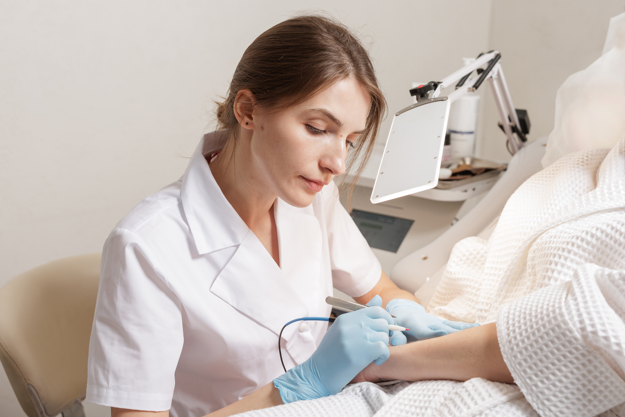 Female specialist performing electrolysis hair removal on female hand using fine probe while patient rests on treatment chair in a clinical cosmetology room with soft indoor lighting.

