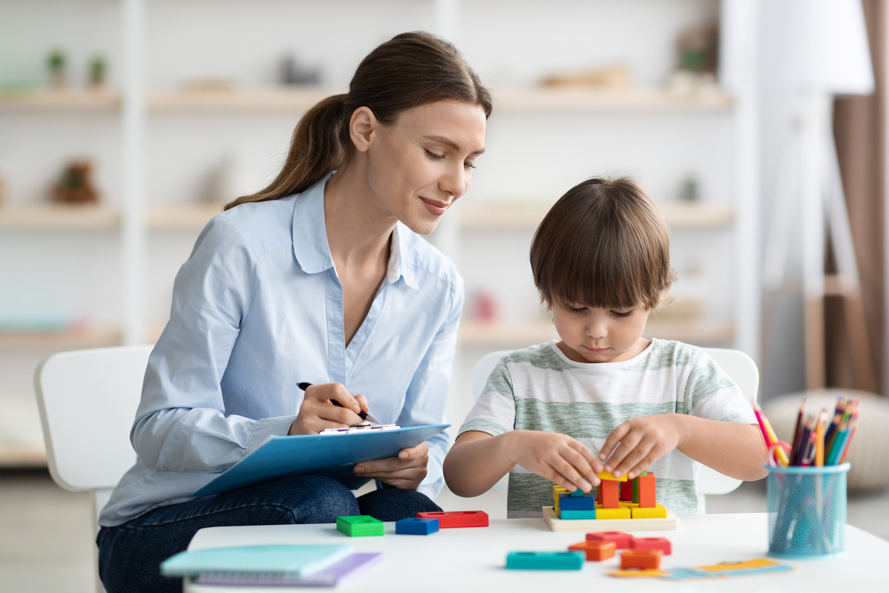 Assessment of kids mental development. Professional woman watching little boy playing with logical game, evaluating his behavior. 

