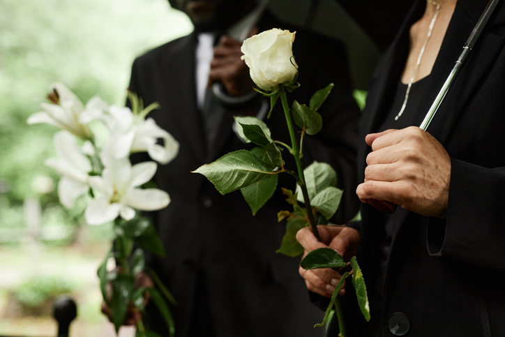 Close up on hands with white rose of unrecognizable senior woman grieving during memorial service at cemetery before placing flowers on grave to commemorate the deceased. 

