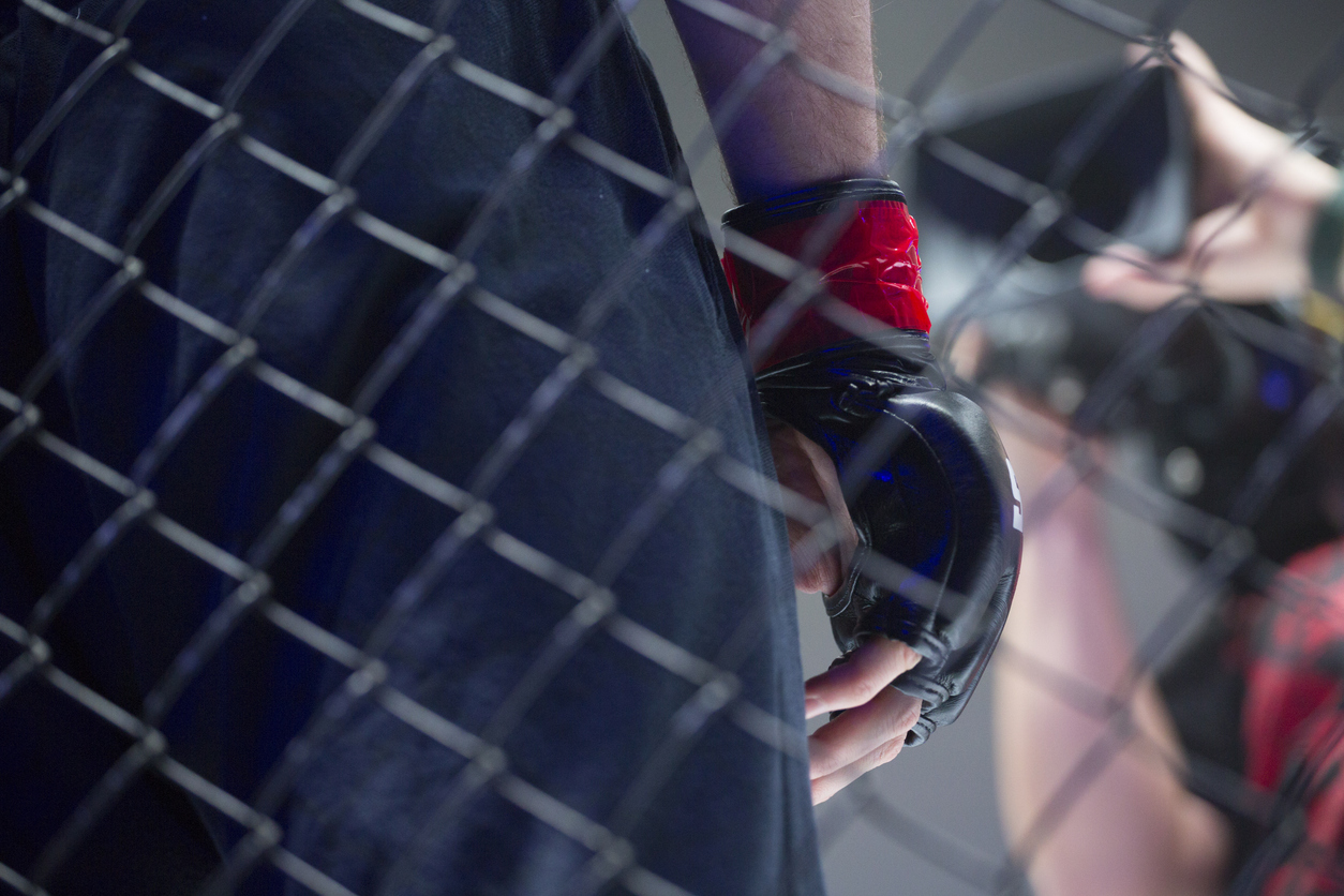 Close up hand in martial arts glove behind a metal wire cage. The background suggests a fight arena.