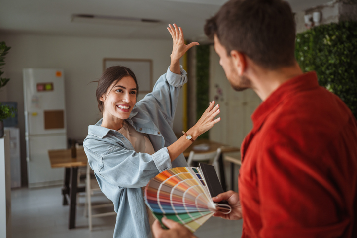 Smiling woman pointing at wall while choosing paint color with interior designer holding a color palette, discussing home renovation project.