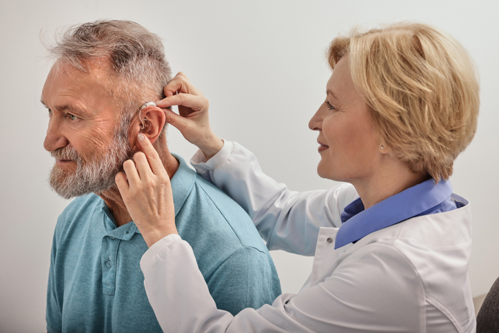 Experienced female doctor fitting Behind-The-Ear hearing aid on senior patient's ear while in an audiology clinic.